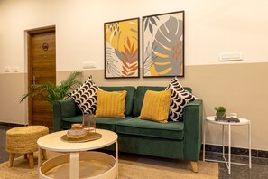 A green sofa with patterned cushions, a small round table with a candle, and a potted plant. - Olive Zip Domlur by Embassy Group (Bengaluru)