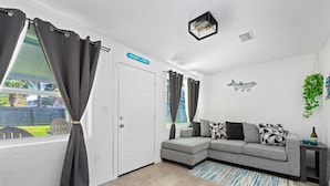 A modern living room with a grey sectional sofa, a small side table, a decorative fish wall hanging, and a view of the outdoors through a window with grey curtains.