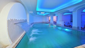 Ein moderner, überdachter Pool mit blauem Licht, klarem Wasser und Liegestühlen am Rand.