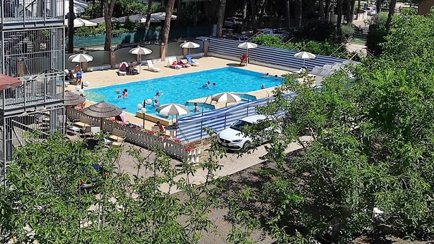 A swimming pool with lounge chairs and umbrellas, surrounded by trees and buildings. - Residence Quadrifoglio (Cervia)