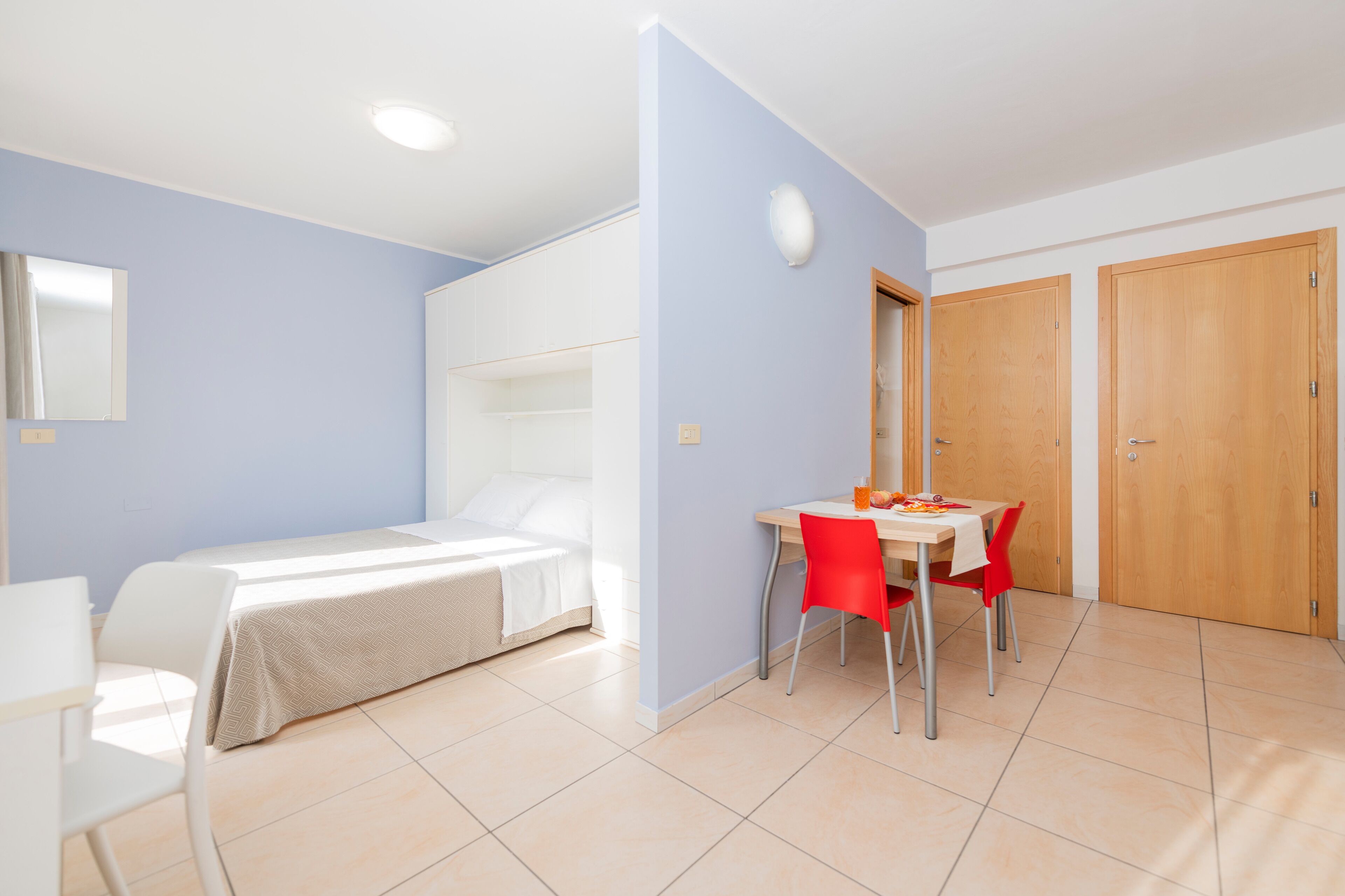 Ein Zimmer mit einem Bett, einem Schreibtisch und einem Stuhl. Eine Holztür und ein Spiegel sind an der Wand angebracht.