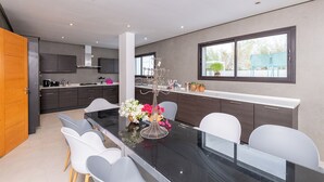 A modern kitchen with dark cabinets, a white countertop, and a dining area with a glass table and white chairs. - Villa Tanit  (La Marsa)