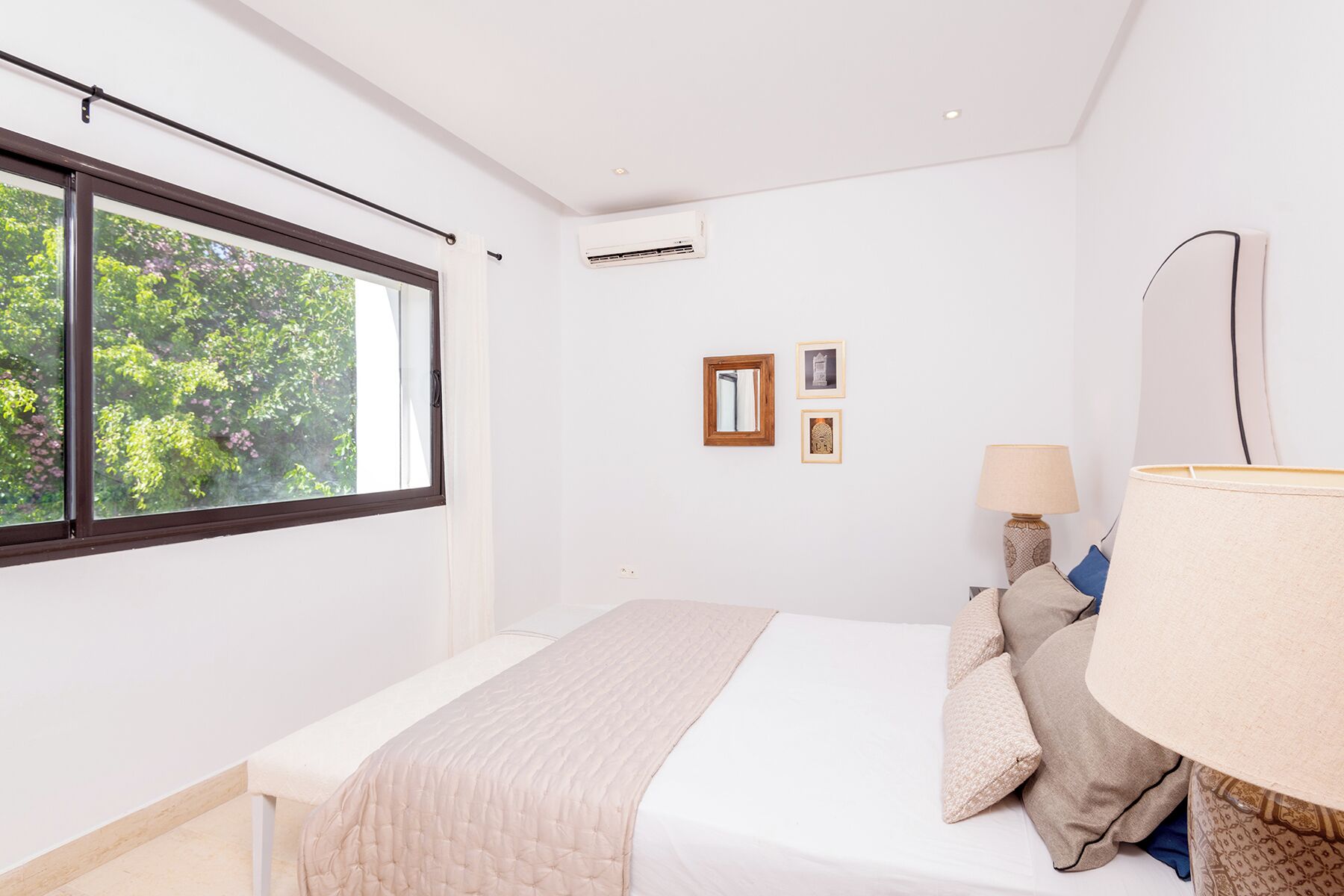 Een slaapkamer met een groot raam, een bed met een beige deken, twee ingekaderde foto's aan de muur en een bedlampje.