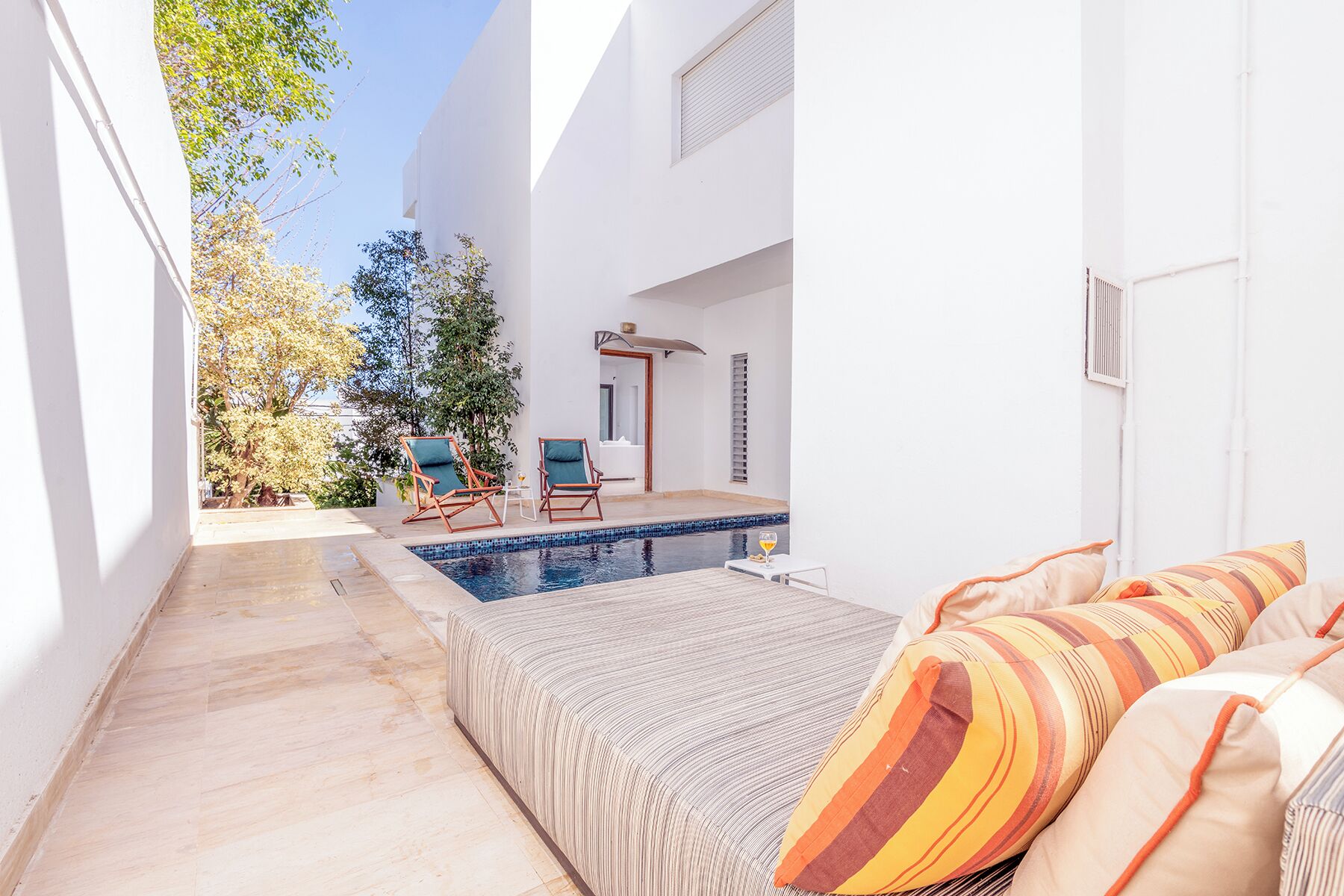 A modern outdoor pool area with a lounge chair and a small table, a covered seating area, and a bed with striped bedding.