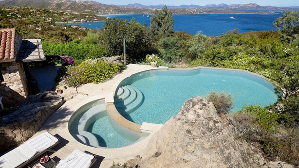 A pool with a curved edge, surrounded by greenery and overlooking a coastal landscape with mountains in the distance. - VILLA LILIUM (Punta Molara)