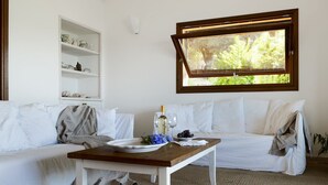 A white sofa with a wooden table in front, a window with a view of greenery, and a white shelf with decorative items. - VILLA LILIUM (Punta Molara)