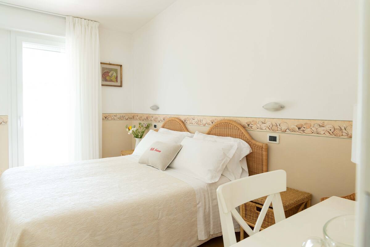 Un lit bien fait, avec du linge de lit blanc, une tête de lit en osier et une petite table de chevet sur laquelle se trouve un verre.