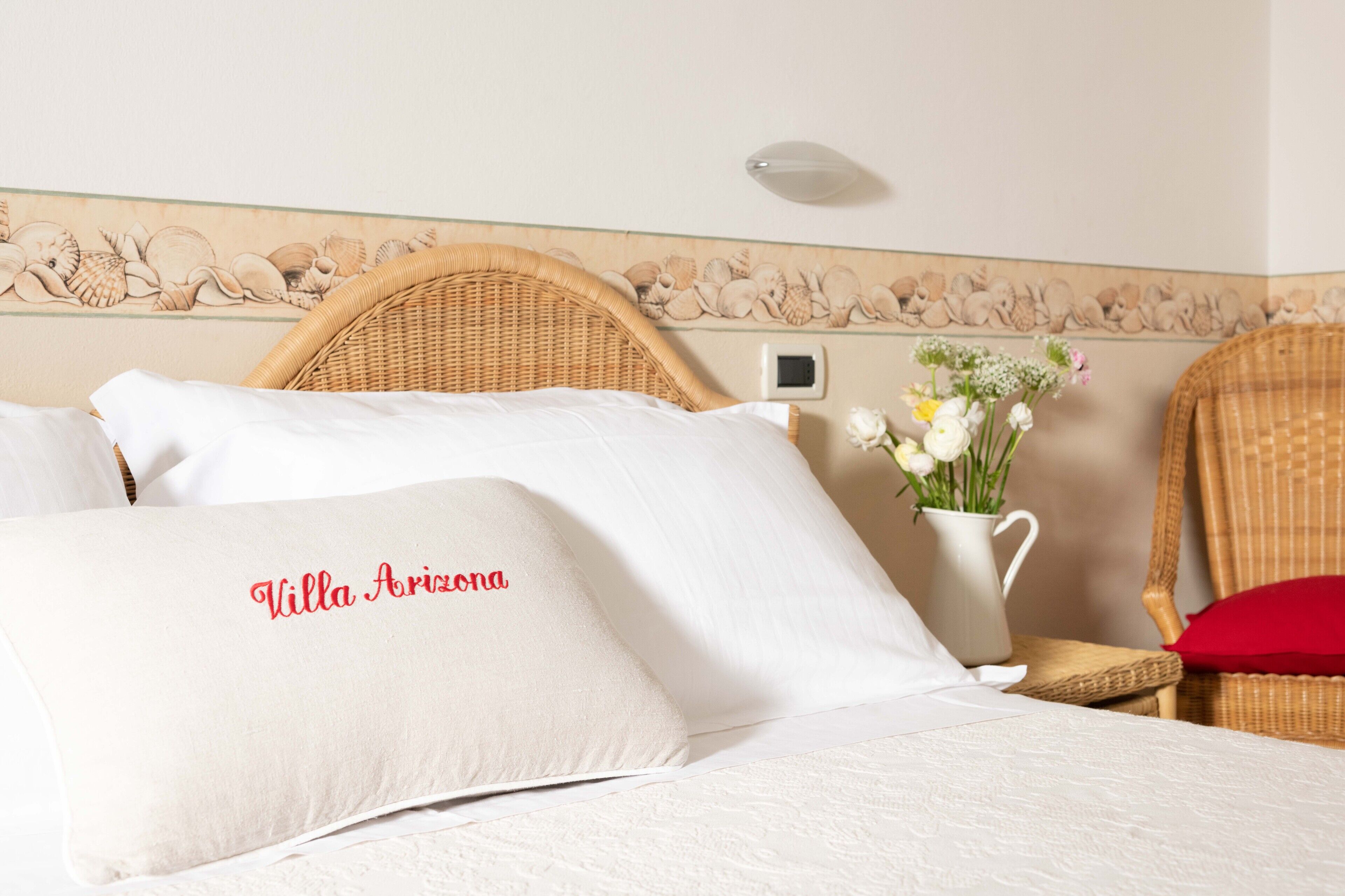 Una habitación de hotel con una cama, una silla de mimbre, un jarrón con flores y un borde de conchas decorando la pared.