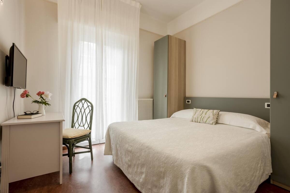Une chambre à coucher avec un lit, un bureau, une chaise, une télévision et une fenêtre avec des rideaux.