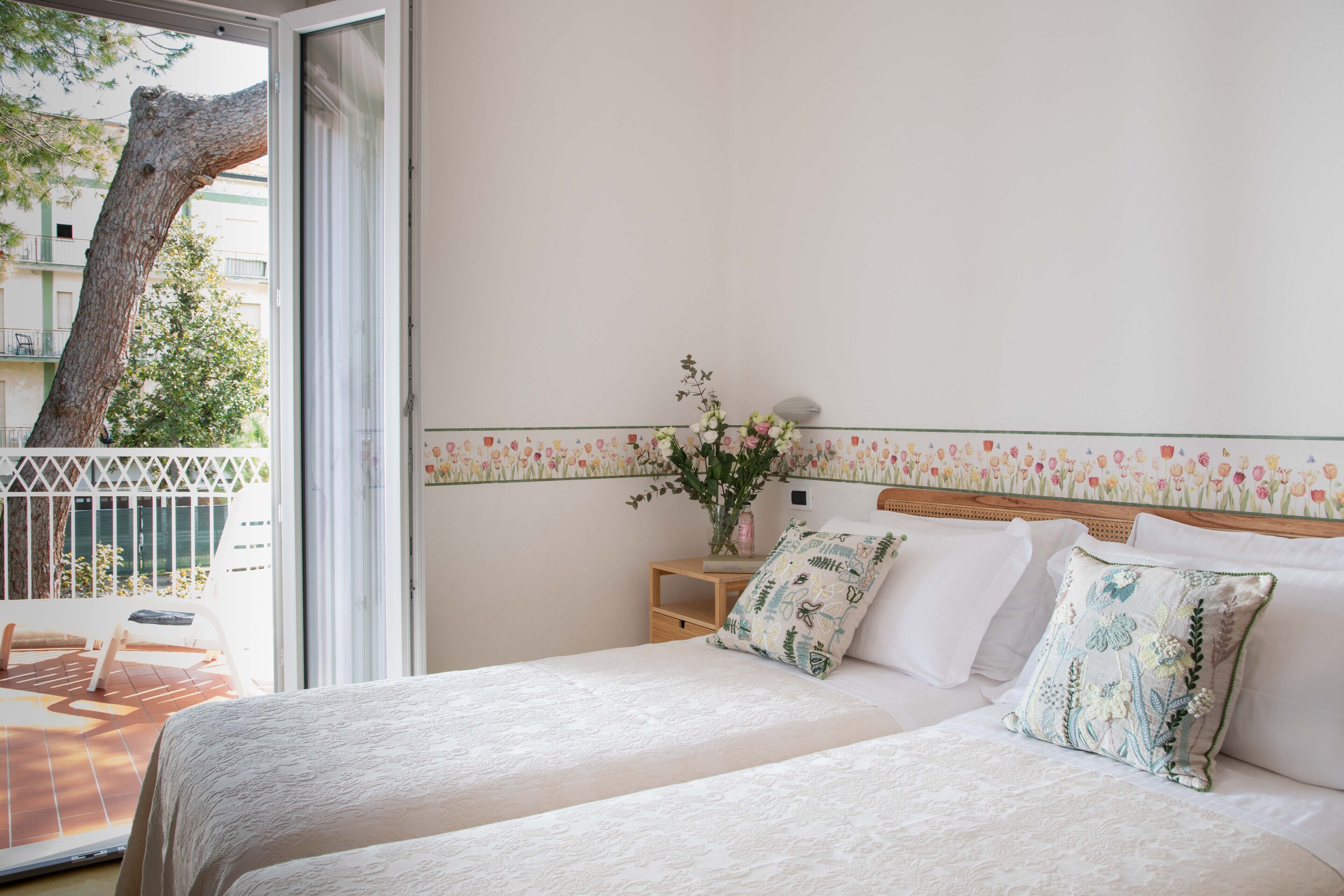 A bedroom with two beds, a nightstand, a vase with flowers, and a balcony with a chair.