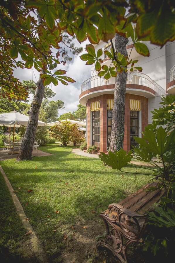 A building with a brick facade, large windows, and a circular balcony. There is a grassy lawn with a bench and umbrellas. - Villa Arizona B&B (Cervia)