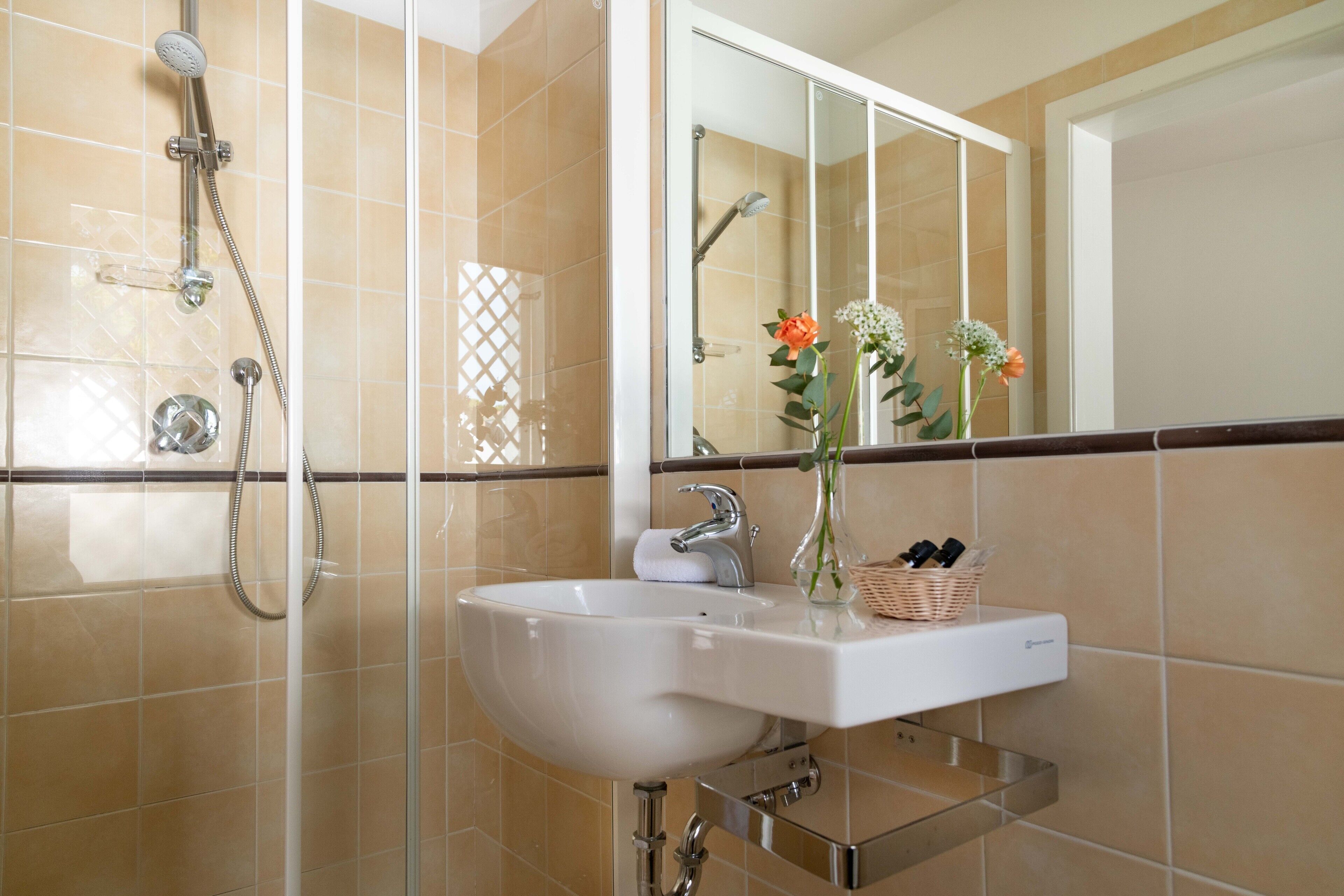 A bathroom with a white sink, a mirror, a shower, and a vase with flowers.