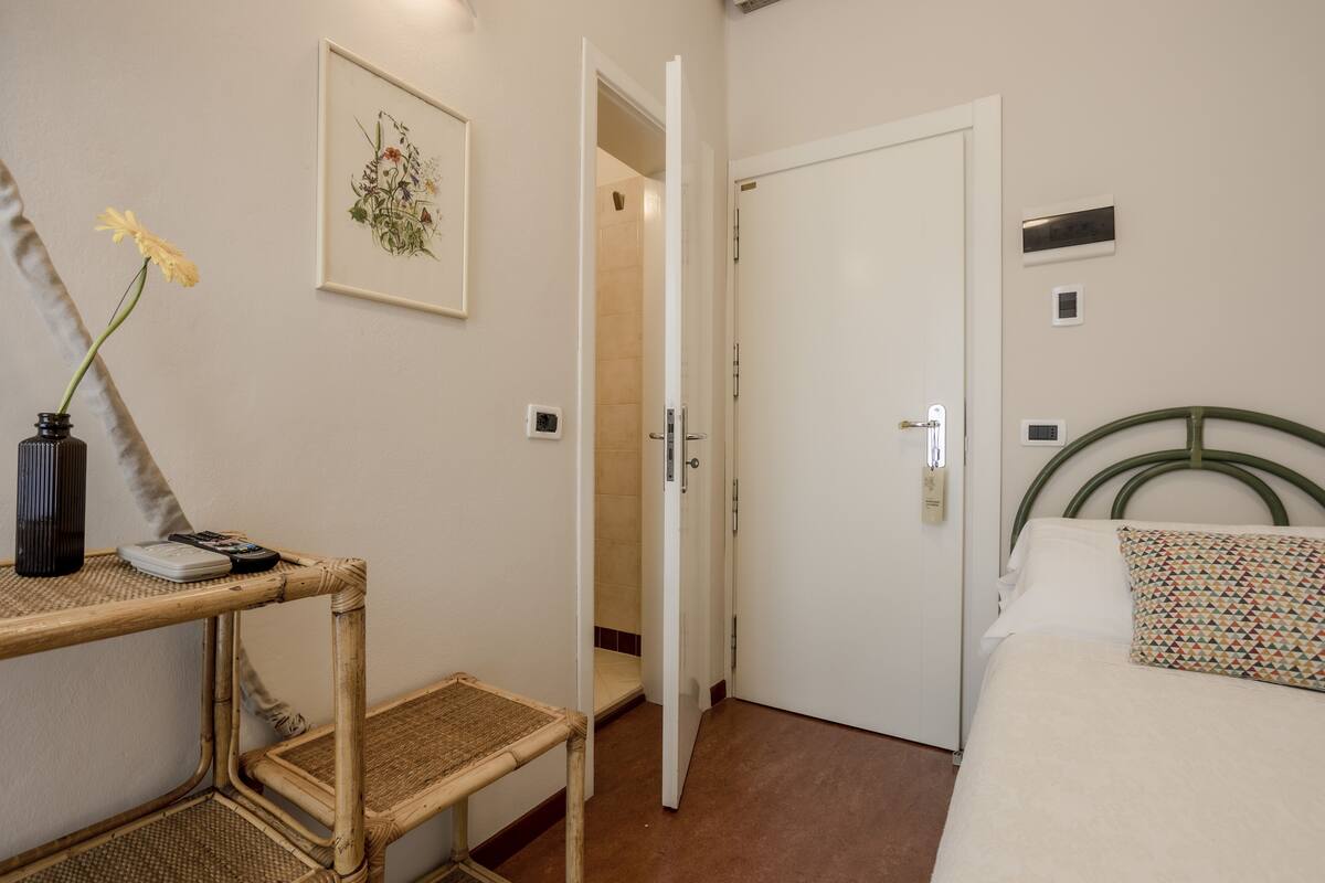 Une pièce comprenant un lit, une table de chevet avec un vase et une télécommande, une porte donnant sur une autre pièce et un interrupteur fixé au mur.