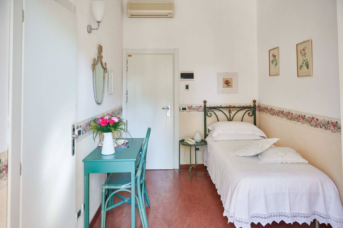 Une petite pièce propre, avec un lit, un bureau et une chaise, une petite table ornée d’un vase de fleurs et un miroir fixé au mur.