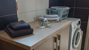 A laundry area with a washing machine, stacked towels, and a laundry basket. - LES Sables (Yves)