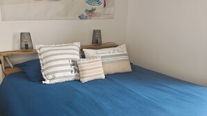 A bedroom with a blue bedspread, striped pillows, and a beach-themed wall art. - LES Sables (Yves)