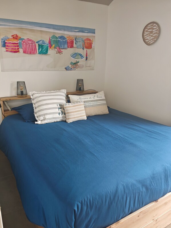 A bedroom with a blue bedspread, striped pillows, and a beach-themed wall art.