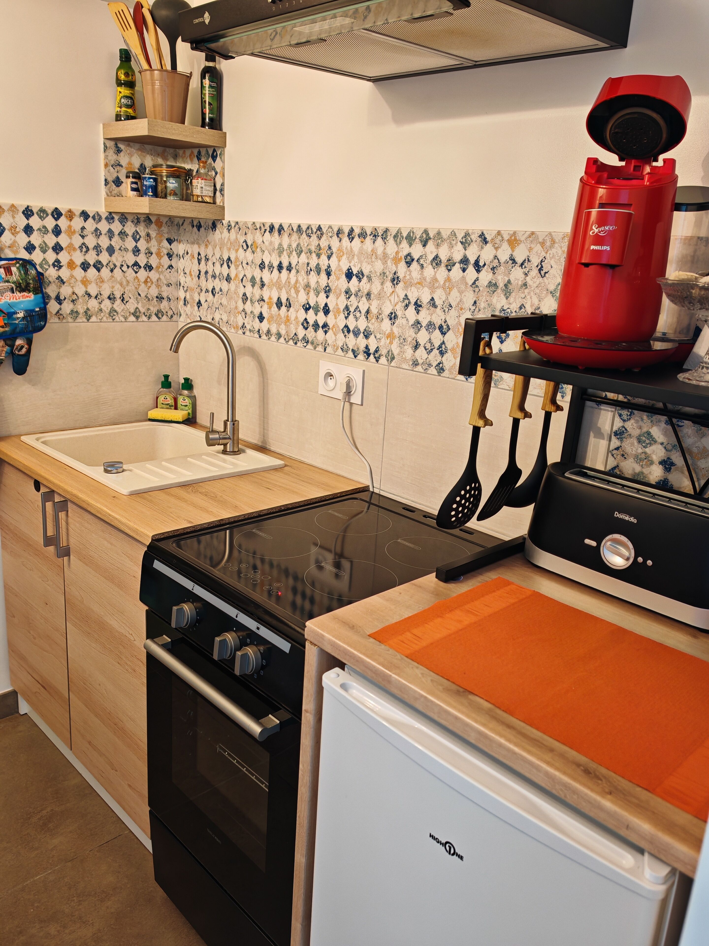 Cocina compacta con lavabo, estufa y refrigerador. Hay una alacena con utensilios de cocina y un electrodoméstico rojo en la encimera.