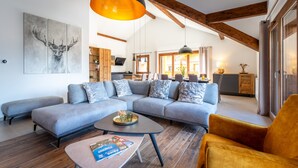 A modern living room with a sectional sofa, a coffee table, and a yellow armchair. There is a painting of a deer on the wall, wooden beams on the ceiling, and a kitchen in the background. - Appartement Ouréa 8 pers navette stations à 100m (Saint-Jean-de-Sixt)