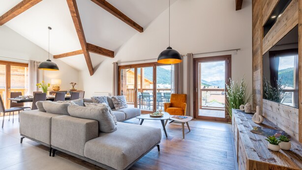 A spacious living room with a grey sofa, wooden beams, large windows, and a wooden reception desk. - Appartement Ouréa 8 pers navette stations à 100m (Saint-Jean-de-Sixt)