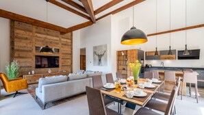 A modern living room with a sofa, dining table, and chairs, wooden beams on the ceiling, and a kitchen area with a TV and wooden cabinets. - Appartement Ouréa 8 pers navette stations à 100m (Saint-Jean-de-Sixt)