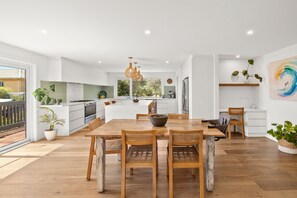 A modern kitchen with white cabinetry, a large island, and a dining area with wooden tables and chairs. - Bliss by the Beach (Aireys Inlet)
