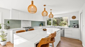 Een moderne keuken met een groot eiland, houten stoelen, hanglampen en uitzicht op groen.