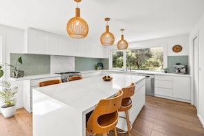 A modern kitchen with a large island, wooden chairs, pendant lights, and a view of greenery outside. - Bliss by the Beach (Aireys Inlet)