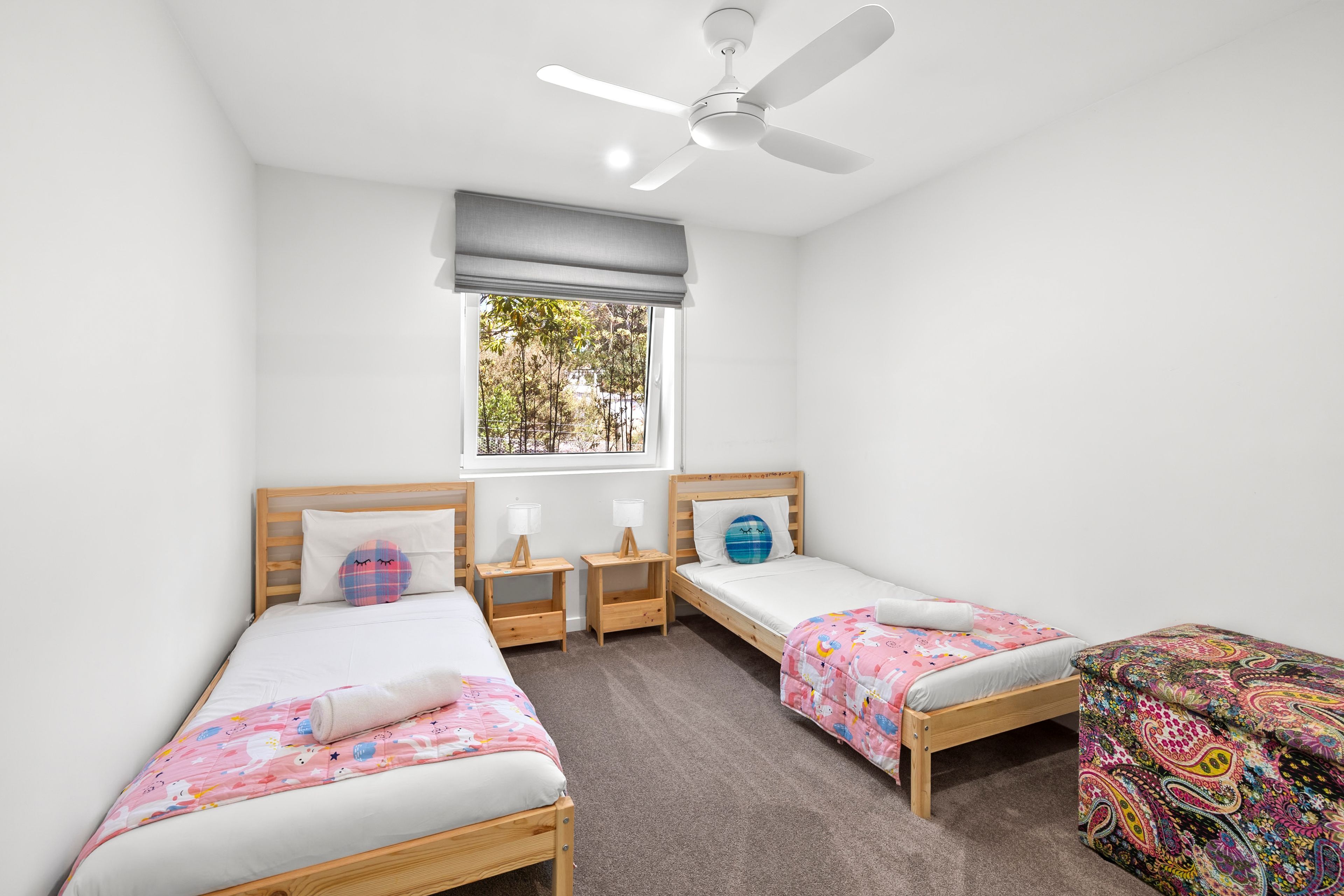 A room with two single beds, a ceiling fan, a window with a view of trees, and a small table with a lamp.