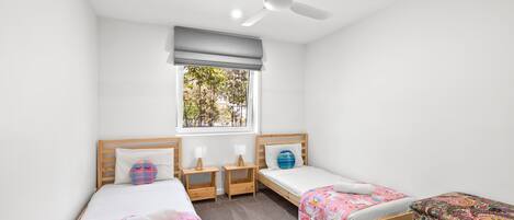 A room with two single beds, a ceiling fan, a window with a view of trees, and a small table with a lamp.