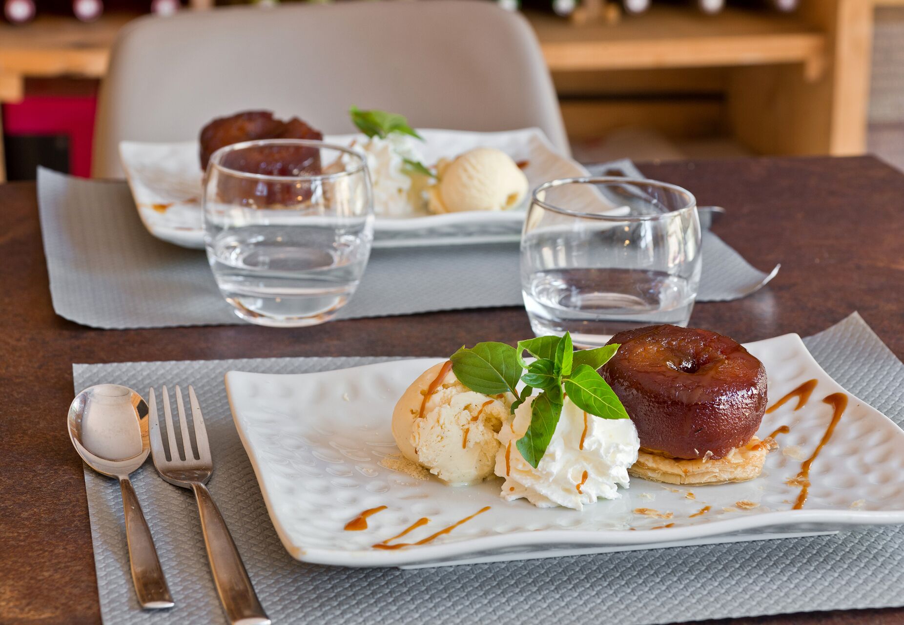 Une assiette à dessert avec une boule de glace et un fruit caramélisé, le tout présenté sur une assiette blanche carrée, accompagné de deux verres d’eau sur une table.