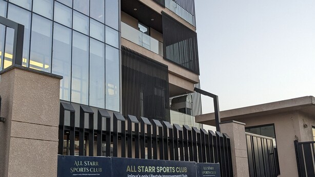 A modern building with a glass facade and a sign for All Starr Sports Club, indicating amenities like a swimming pool, table tennis, billiards, and a gym. - All Starr Sports (Jaipur)