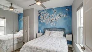 Een slaapkamer met een groot bed, een plafondventilator, een nachtkastje, een dressoir en een muurschildering.