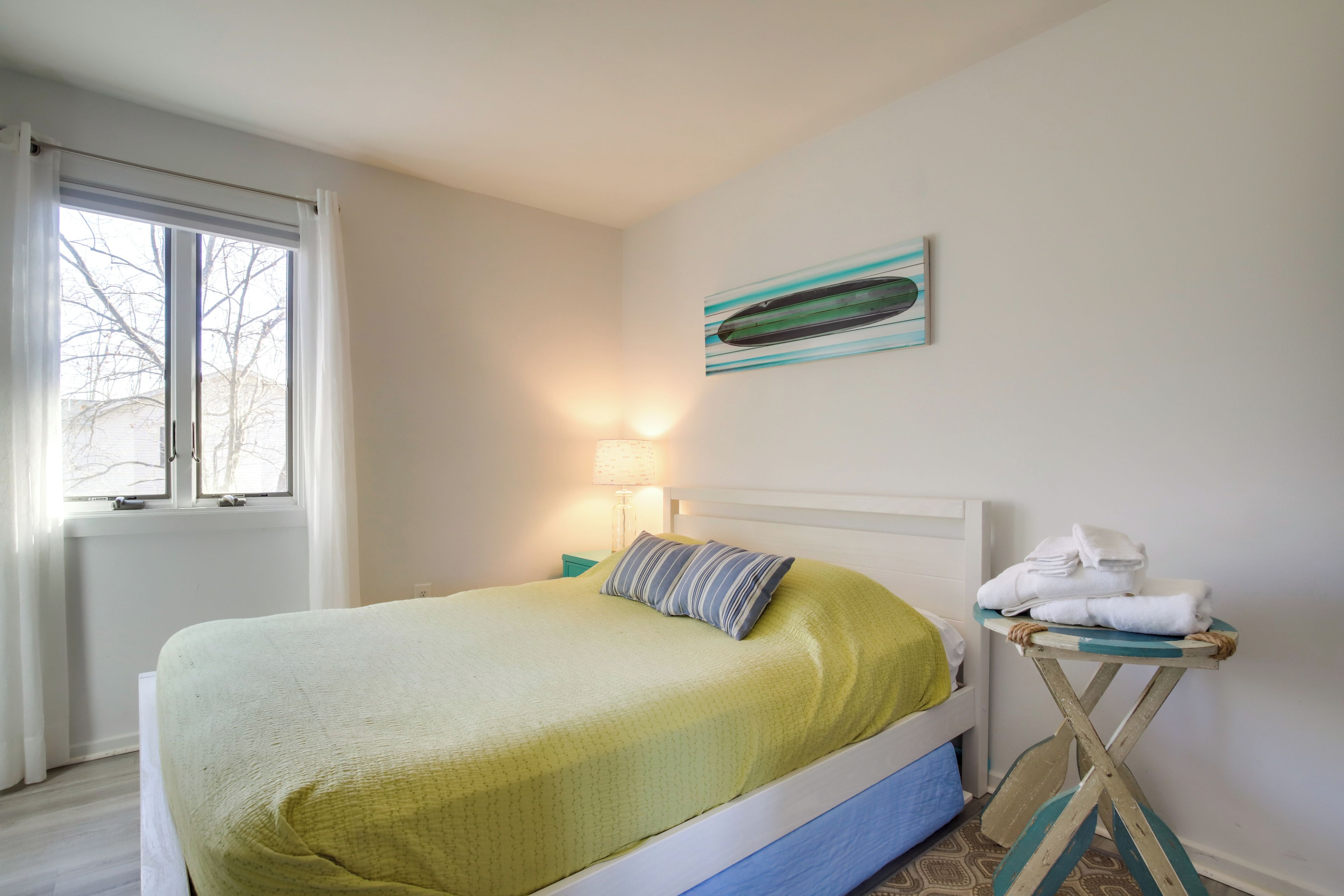 A neatly made bed with a green bedspread and a striped pillow, a small table with towels, a lamp, and a painting on the wall.
