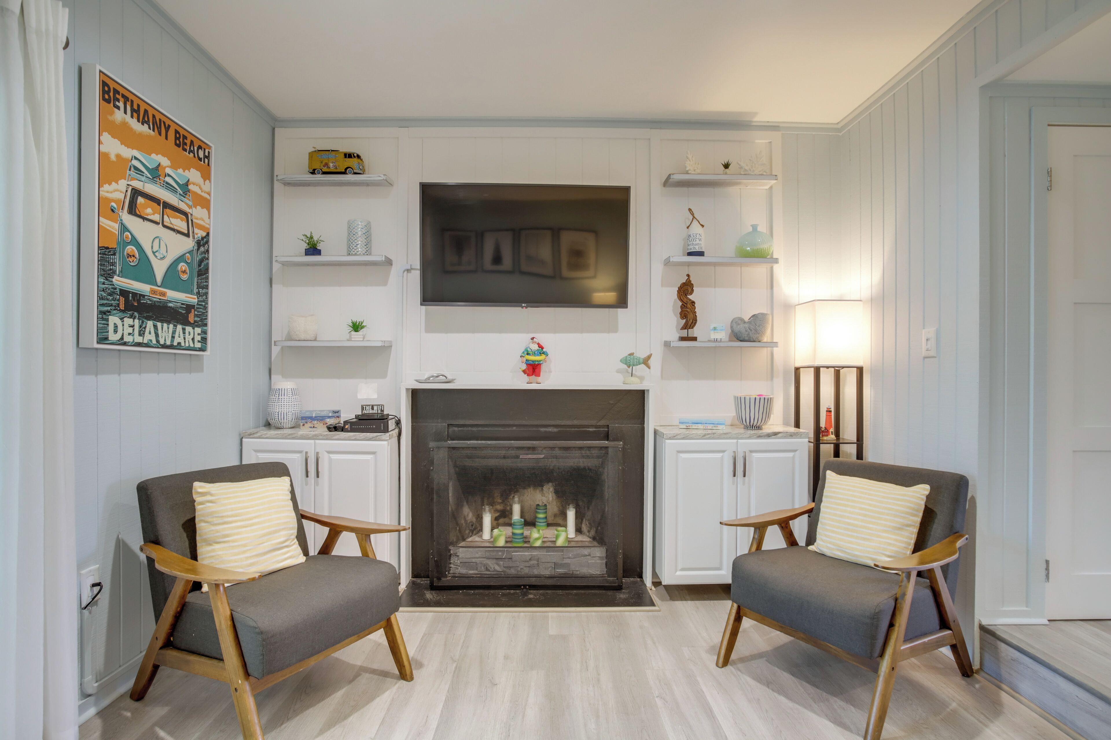 A living room with a fireplace, two armchairs, a TV mounted above the fireplace, and a wall with shelves and a poster.