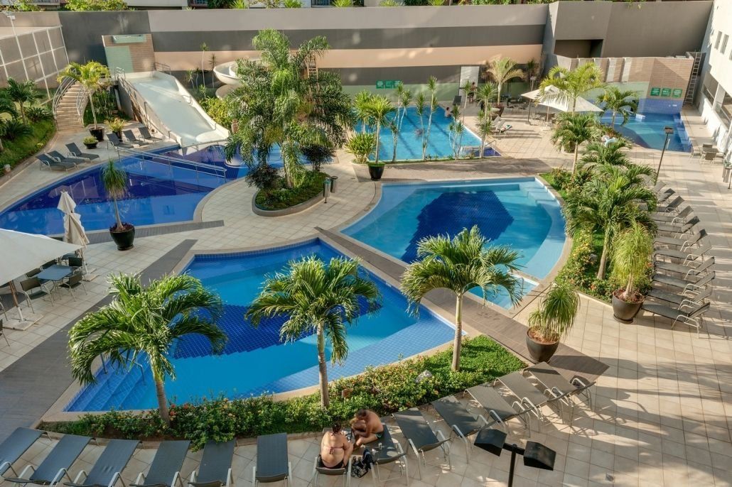 Un área de piscina en la azotea con varias piscinas, palmeras y sillones de descanso.