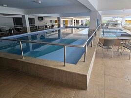 Una piscina interior con barandilla metálica, rodeada de suelo de baldosas y una zona de estar con mesas y sillas.