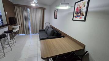 A modern living room with a wooden dining table, black chairs, a black leather sofa, a TV mounted on the wall, and a large window with curtains.