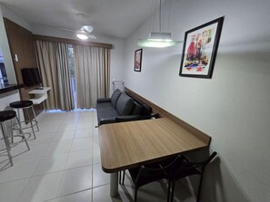 Una sala de estar moderna con una mesa de comedor de madera, sillas negras, un sofá de cuero negro, un televisor montado en la pared y un amplio ventanal con cortinas.