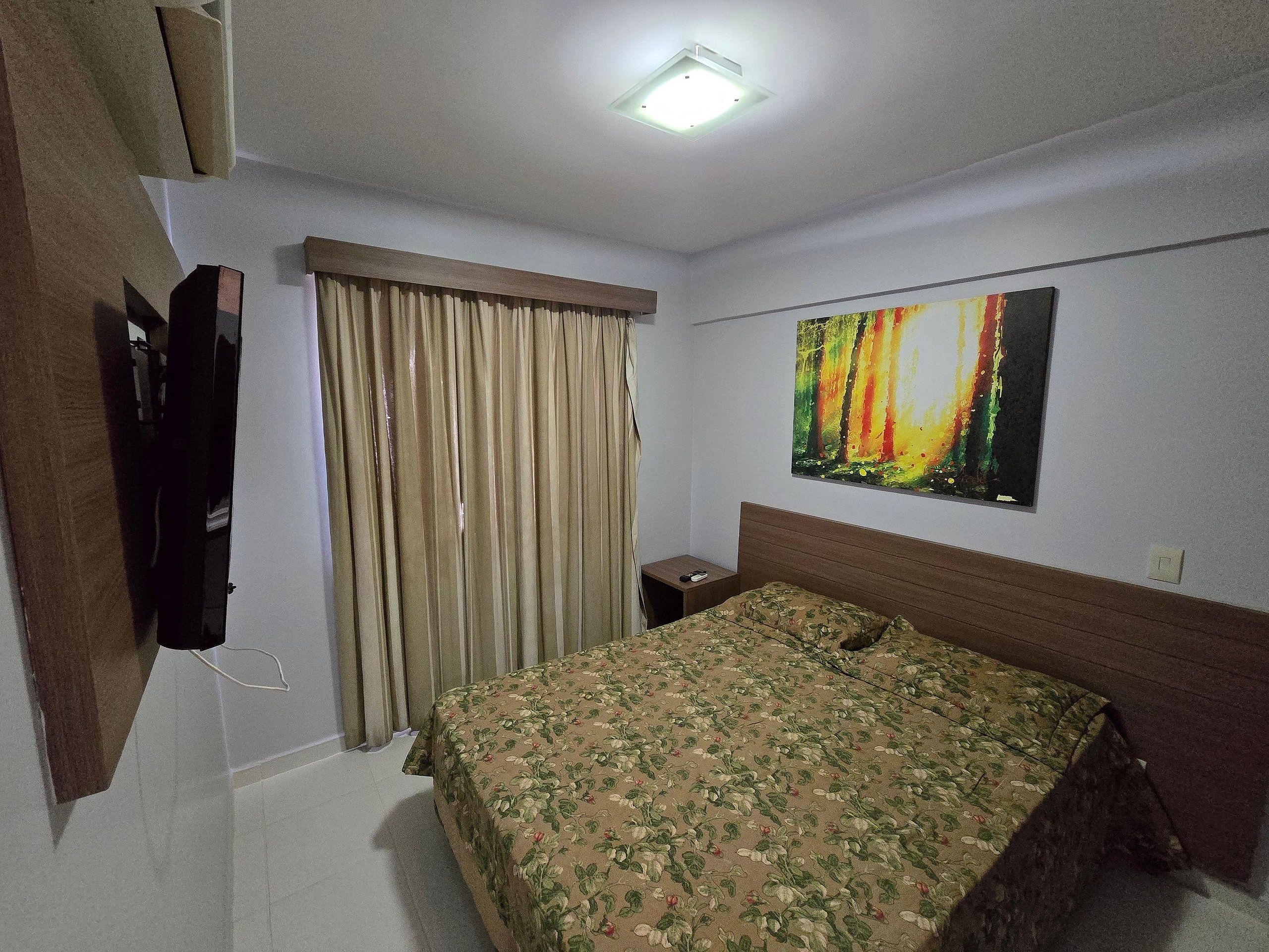 A hotel room with a bed, curtains, a painting, a TV, and a bedside table.