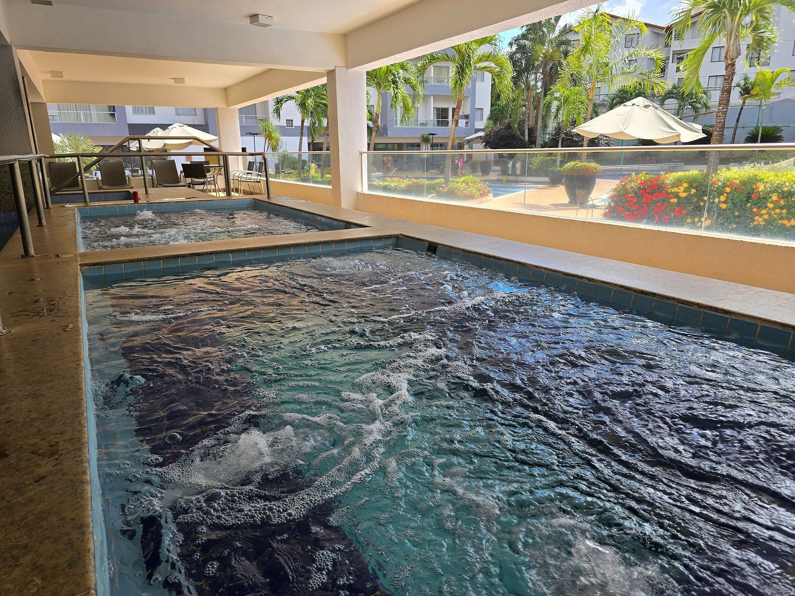 Uma piscina interior ampla com água cristalina, rodeada por uma varanda com assentos e vista para um edifício com palmeiras.