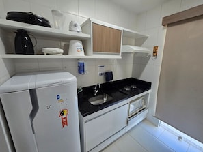 A compact kitchen with white cabinets, a black countertop, a built-in microwave, and a small sink. - Aptos Rio Quente - ótima área de lazer (Rio Quente)