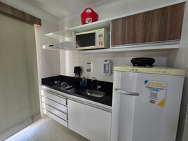 Uma cozinha compacta com fogão a gás, micro-ondas e frigorífico. Há uma pequena prateleira acima do micro-ondas e uma mala vermelha em cima.