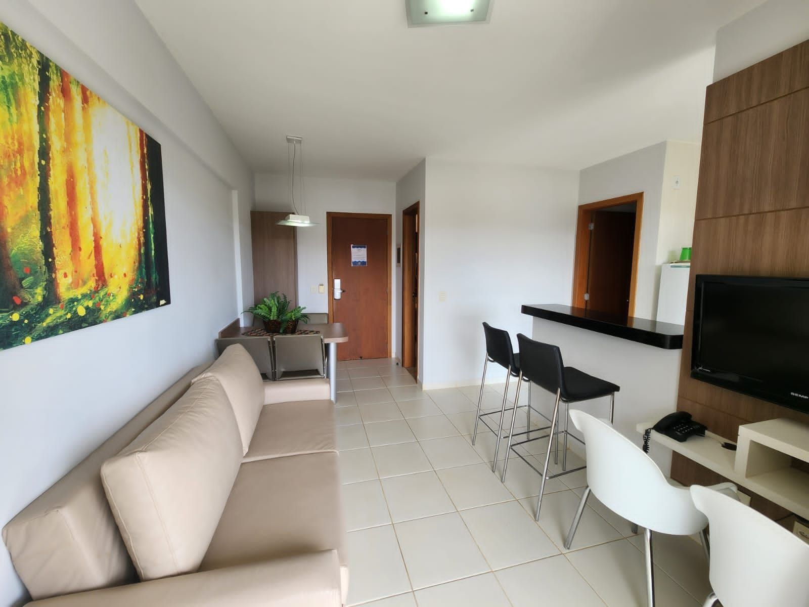 A modern living room with a beige sofa, a dining area with a small table and chairs, a kitchen area with a TV, and a large abstract painting on the wall.