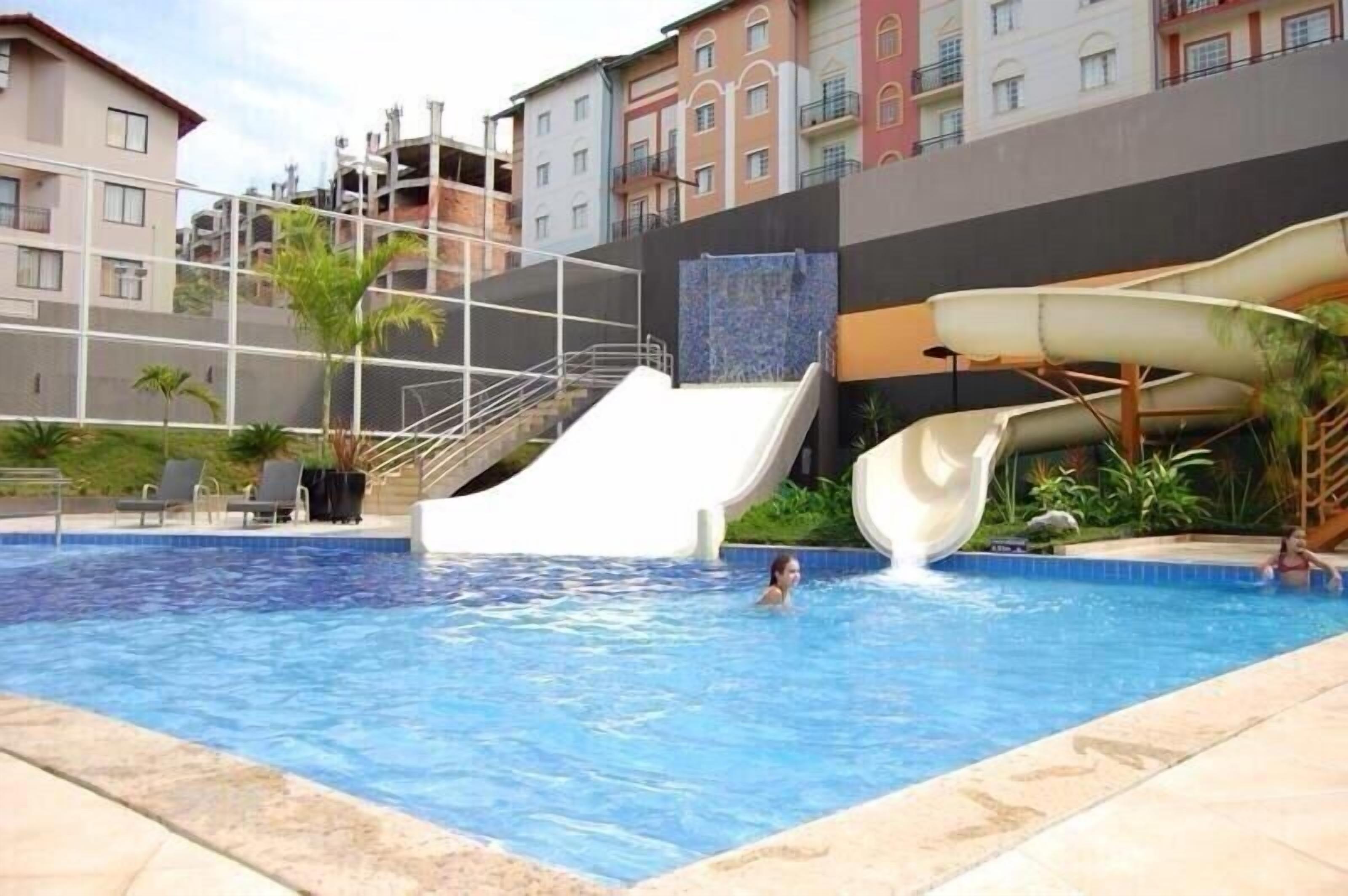 Una piscina con tobogán de agua, rodeada por un edificio con balcones y un cielo despejado.