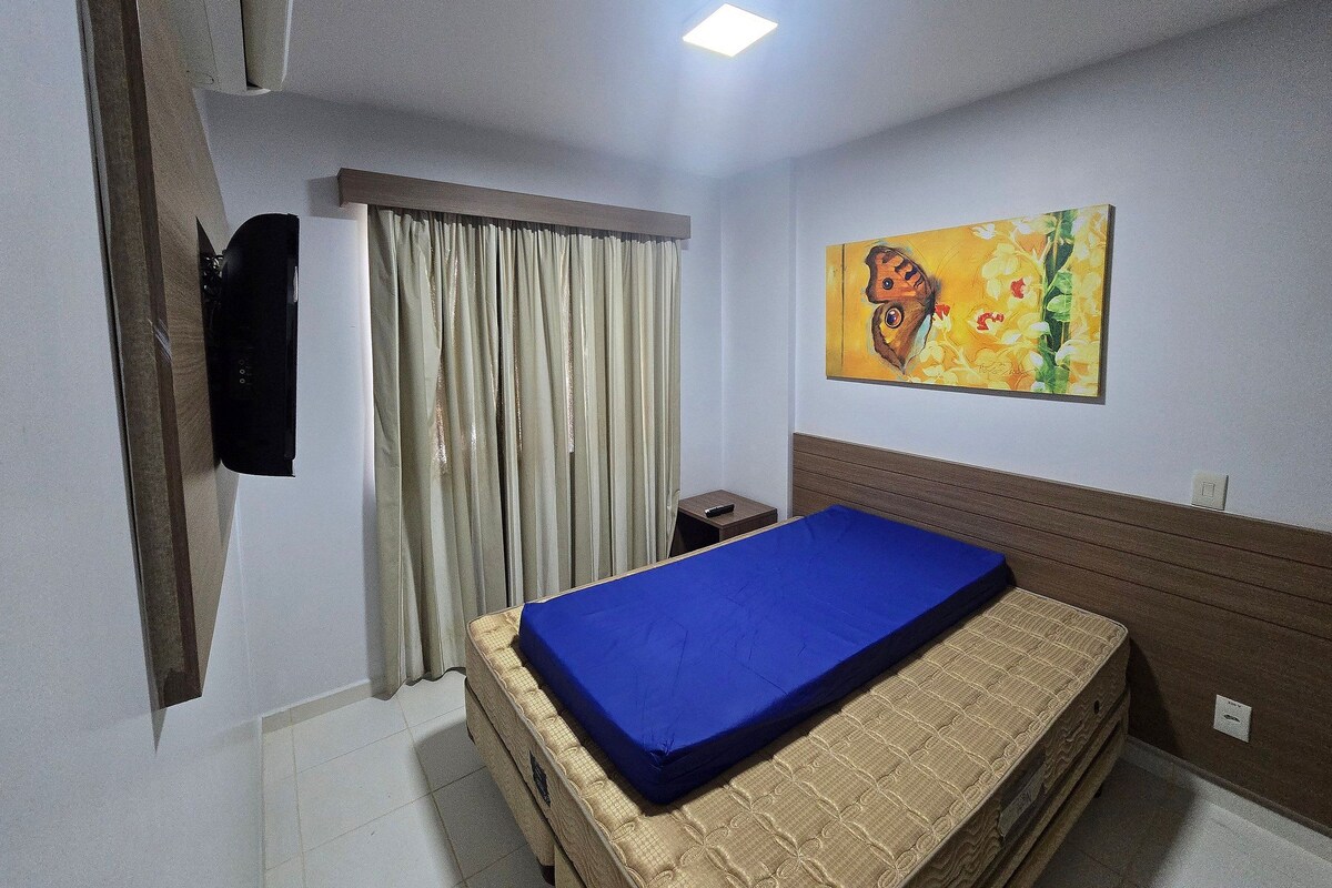 A hotel room with a bed, a painting on the wall, a window with curtains, and a bedside table.