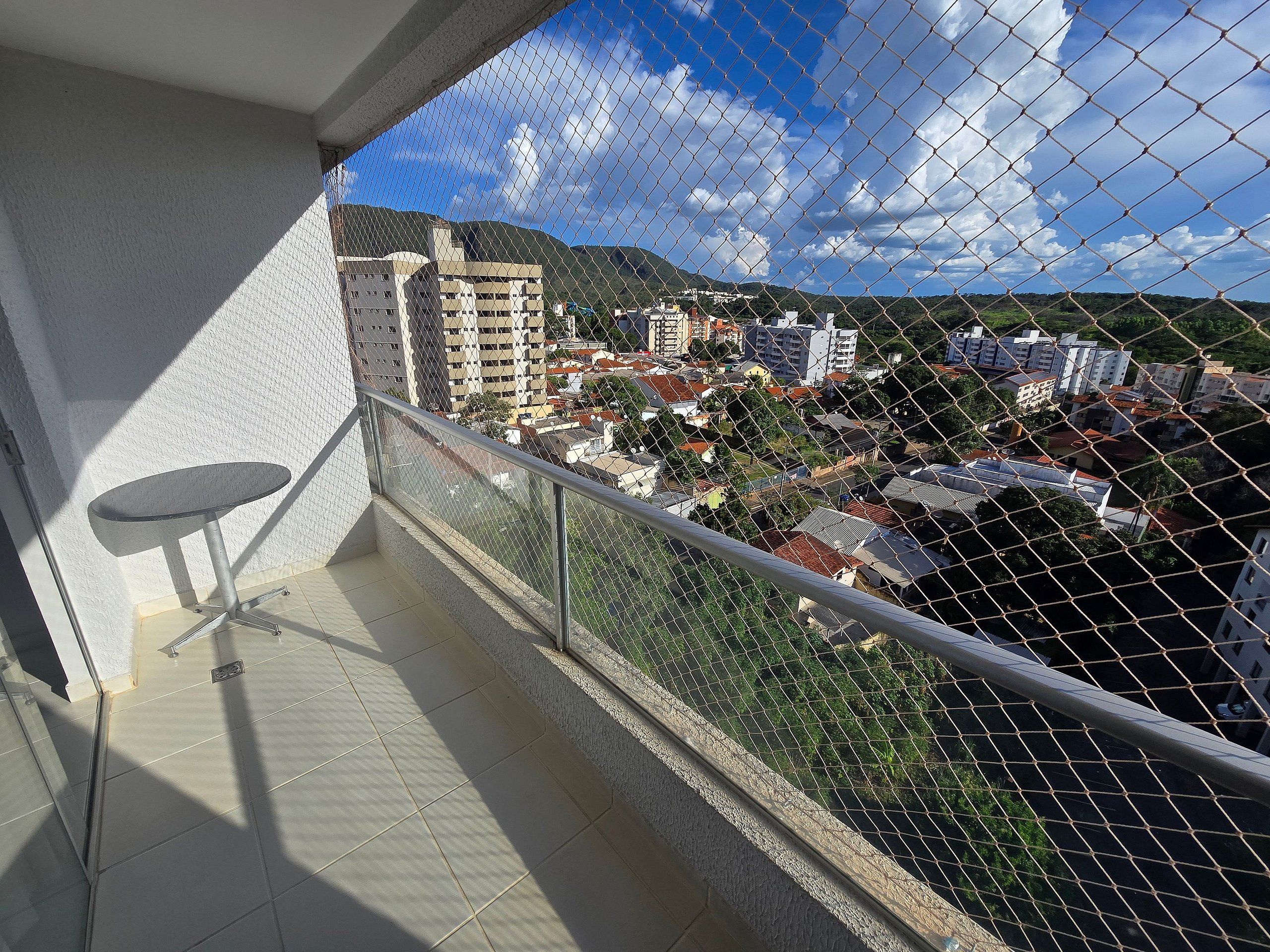 A balcony with a chain-link fence, a small table, and a view of a cityscape with buildings and hills.