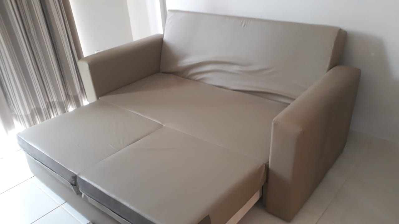Um sofá bege com uma mesa dobrável numa sala com parede branca e janela com cortinas.