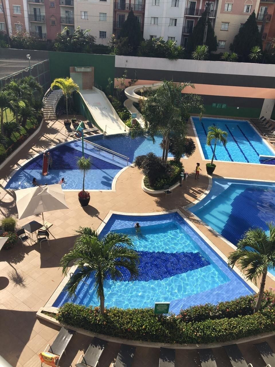 A rooftop swimming pool area with multiple pools, lounge chairs, and palm trees.