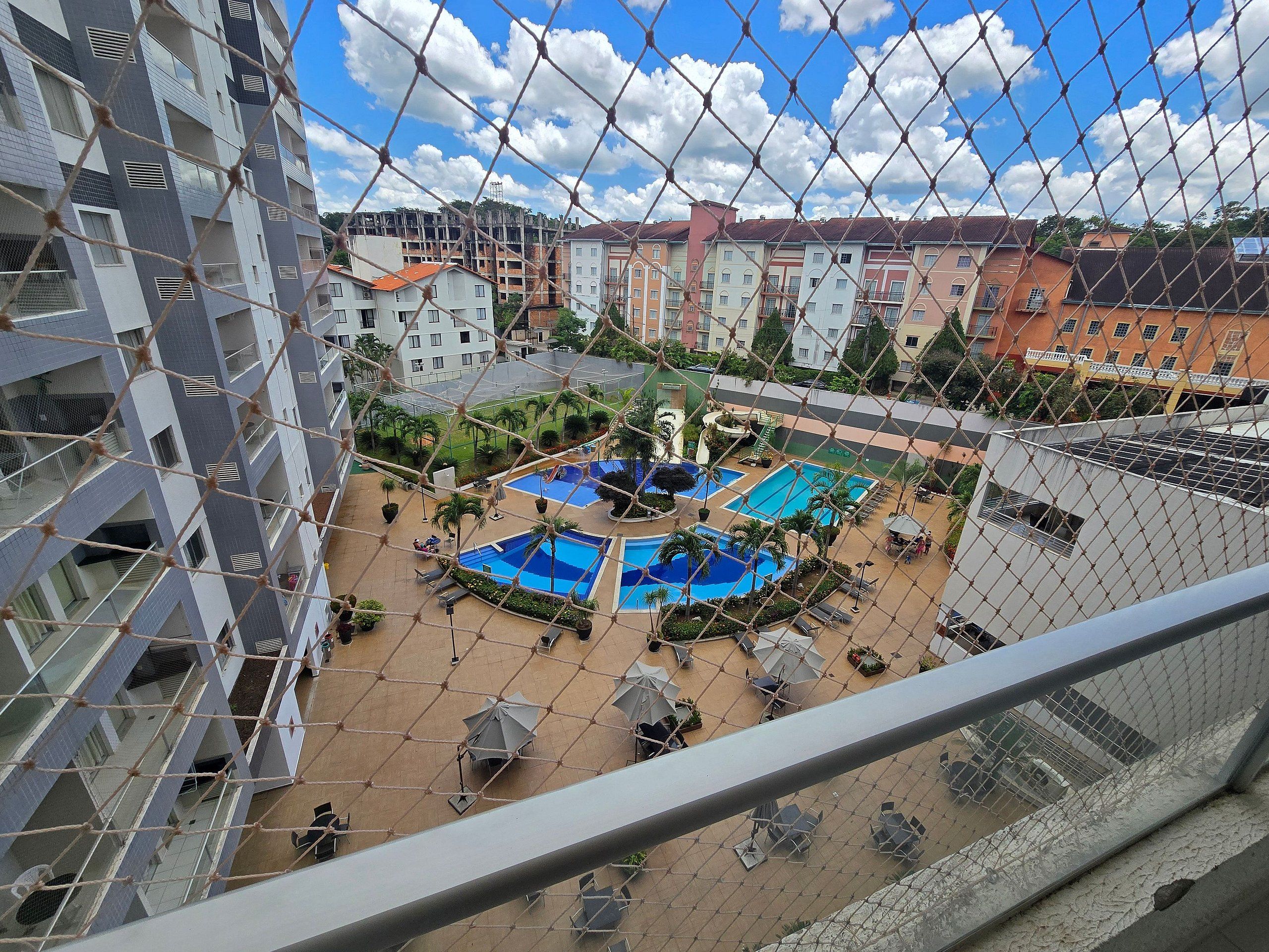 Vista de un edificio residencial con varios balcones, una zona de piscina con tumbonas y sombrillas, y un cielo azul despejado.